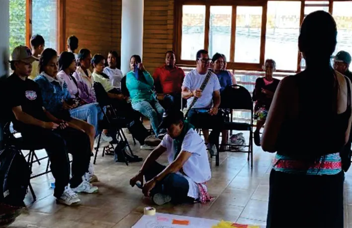 Taller en un salón: personas sentadas en sillas y una mujer de pie con traje tradicional, de espaldas.