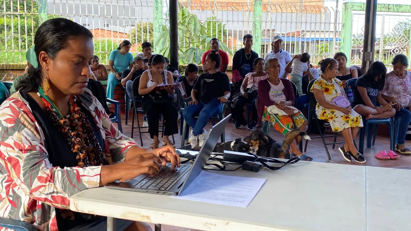 Mujer con collares escribiendo en un computador portátil; detrás, asistentes sentados en un salón abierto.