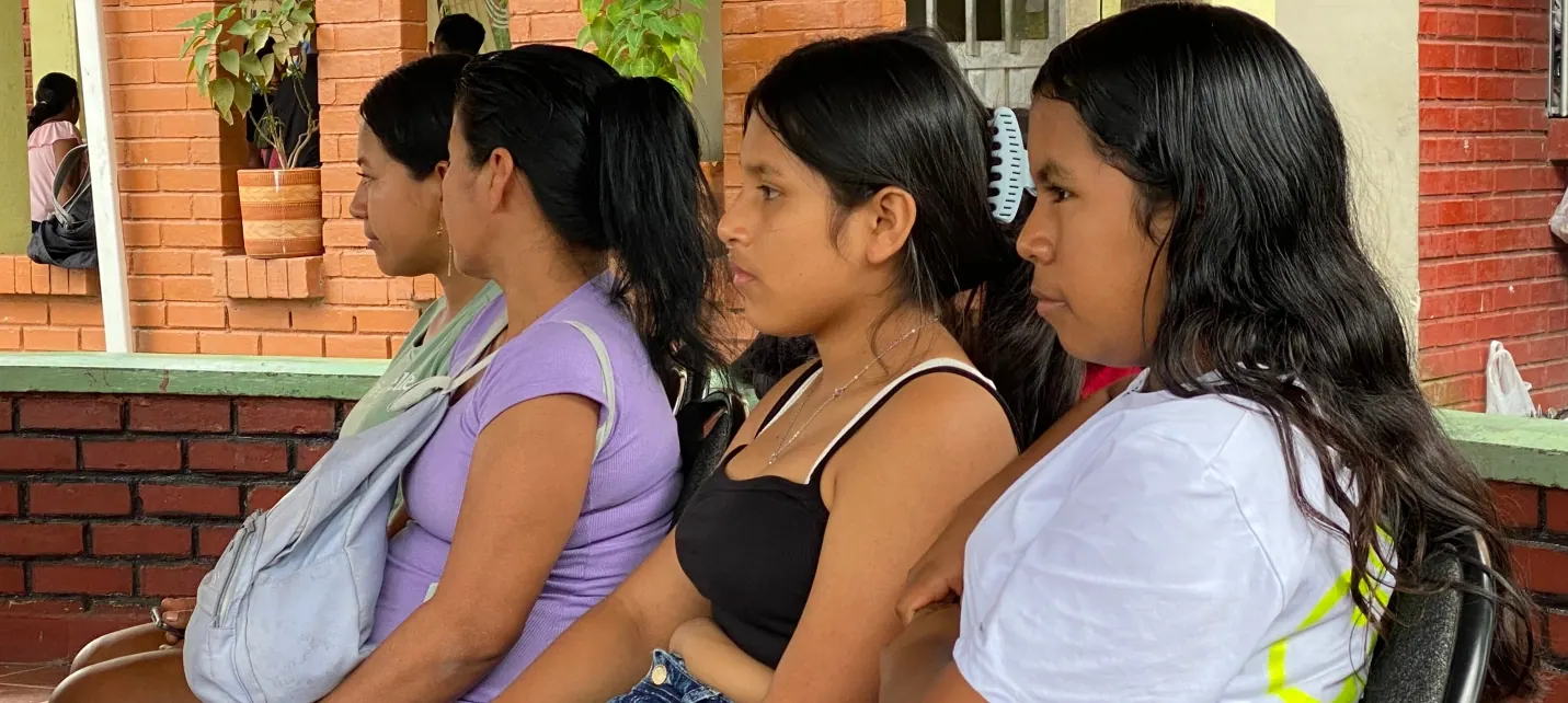 Tres mujeres jóvenes sentadas mirando hacia el frente, junto a una pared de ladrillo.