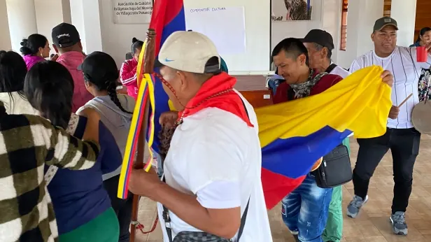 Mayor con pañoleta roja y bastón danzando con una bandera de Colombia en un salón.