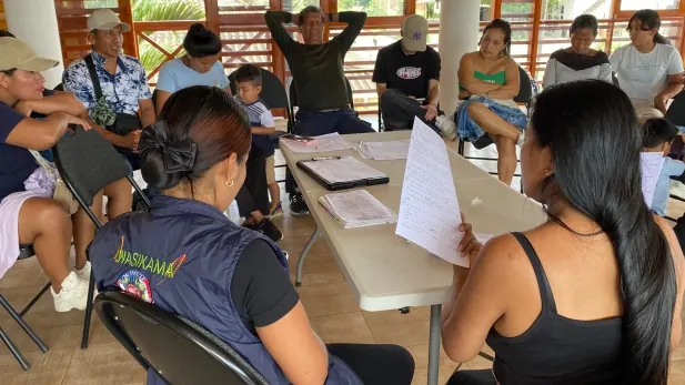 Mujeres revisando documentos en una mesa; asistentes al fondo.