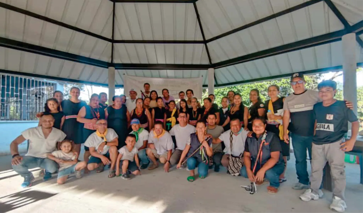 Grupo de comuneros y comuneras de pie y agachados bajo un kiosco.
