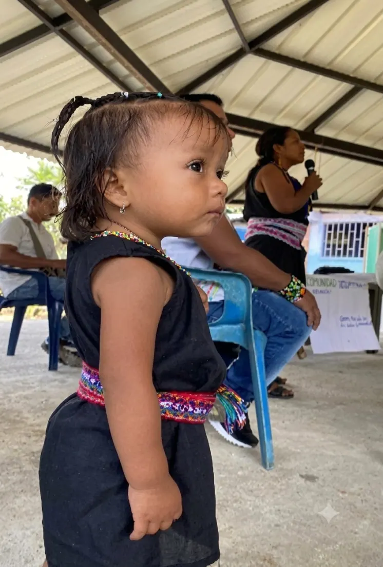 Niña pequeña de pie con vestido negro y faja tejida; al fondo personas en una reunión.