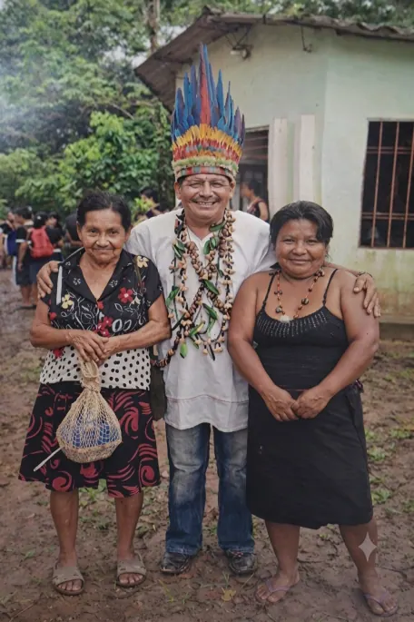 Mayor con corona de plumas y collares entre dos mujeres, frente a una casa.