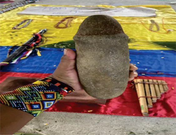 Mano sosteniendo una piedra tallada sobre una bandera, junto a una flauta de caña (otra toma).