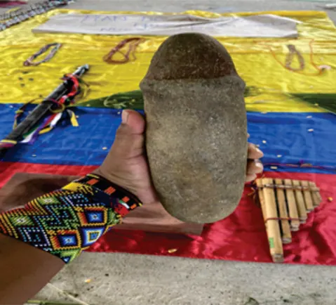 Mano con manilla de chaquira sosteniendo una piedra tallada sobre una bandera, junto a una flauta de caña.