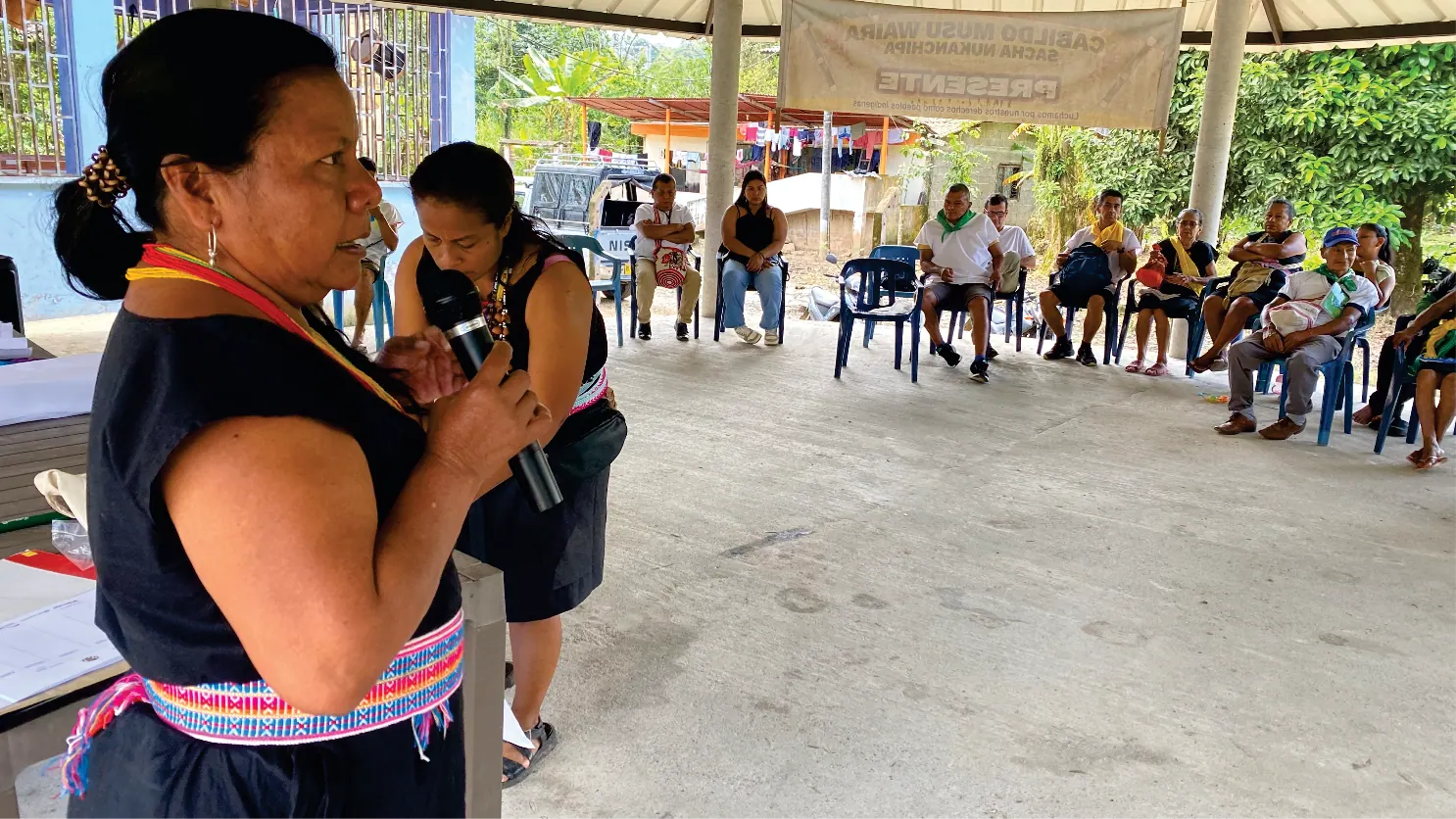 Mujer con traje tradicional hablando con micrófono en una reunión bajo un kiosco.