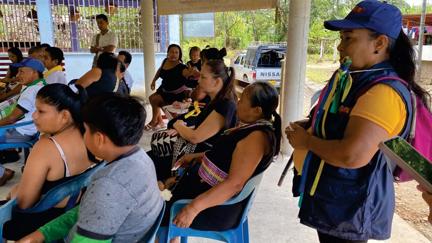 Personas sentadas en una reunión bajo techo; mujer de pie con chaleco.