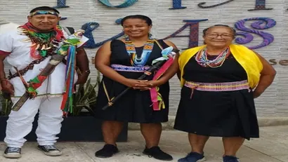 Un hombre y dos mujeres con traje tradicional y collares frente a un letrero.