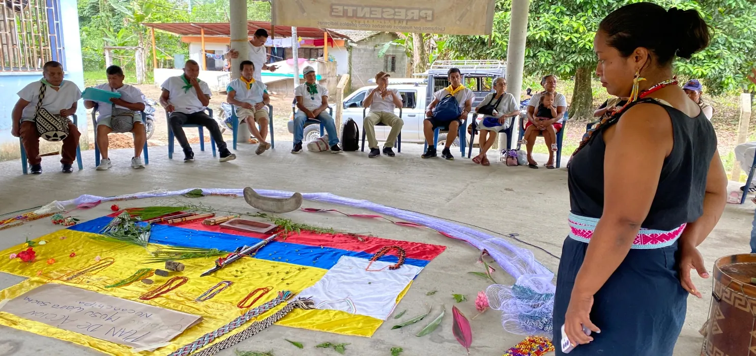 Reunión en círculo con tejidos y banderas extendidos en el suelo; mujer de pie con traje tradicional.