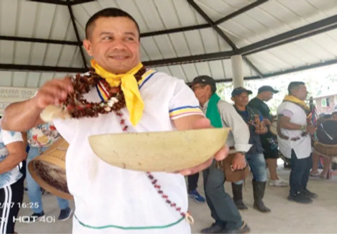Hombre con pañoleta amarilla mostrando una totuma; personas al fondo.