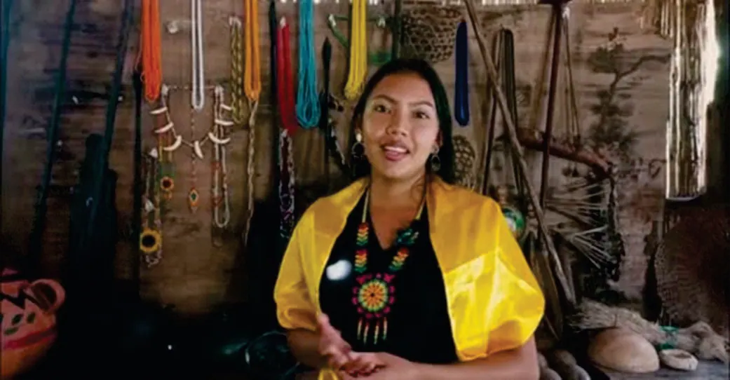 Mujer joven con chalina amarilla y collares sentada en un espacio con artesanías colgadas.