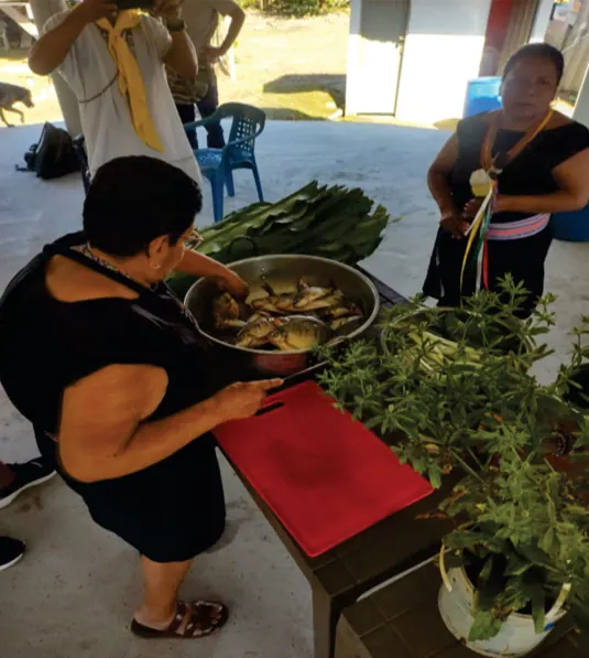 Mujeres preparando alimentos con hojas y una olla.