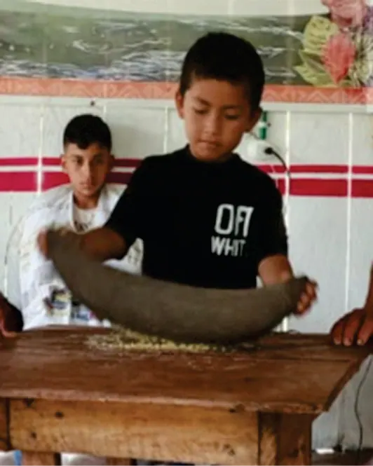 Niño moliendo maíz en una batea de madera.